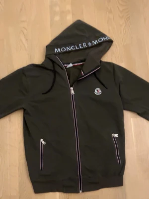 Mörk grön Moncler hoodie med dragkedja - Snygg mörk grön hoodie från Moncler med dragkedja framtill och två fickor med vita detaljer. Huva med snörning och Moncler-logga på bröstet samt tryck på huvan. Perfekt för en chill och stilren look.
