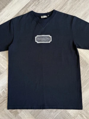 Svart t-shirt  - Stilren svart t-shirt med ett broderat märke på bröstet. Klassisk passform och rund halsringning. Perfekt för dig som gillar exklusiva och tidlösa plagg.
