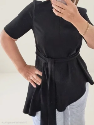 Svart topp från Zara Stl S - Snygg svart topp med rund halsringning och korta ärmar. Toppen har ett långt knytband i midjan som ger en markerad siluett och en lätt utsvängd nederdel. Perfekt till jeans eller kjol för en stilren look.