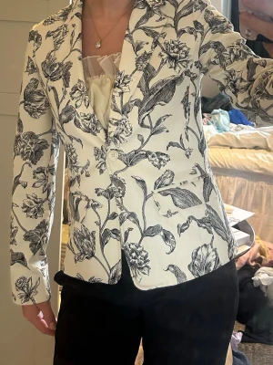 Vit kavaj med svart blommönster - Snygg vit kavaj med coolt svart blommigt mönster. Kavajen har två knappar framtill och figurnära passform. Perfekt för dig som vill sticka ut med en trendig och stilren look. Ger den där extra touchen på din outfit! Jag är en normal S och den sitter bra på mig☺️