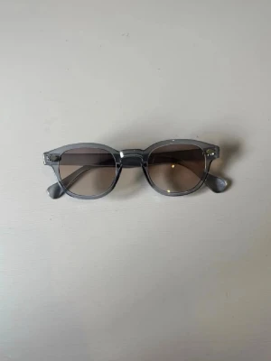 S&T Light Blue Solglasögon  - Snygga solglasögon med transparent grå båge och runda linser med lätt toning. Klassisk och stilren design som passar perfekt till sommaren. Bågarna är i plast och ger en modern vibe.