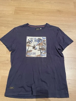 Blå t-shirt med tryck från Morris - Snygg mörkblå t-shirt från Morris med ett stort tryck framtill som visar en strand och palmer. T-shirten har rund hals och korta ärmar. Perfekt för dig som gillar stilrena plagg med coola detaljer.