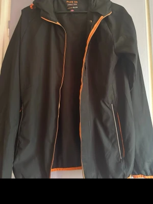 Svart vindjacka med orange detaljer - Snygg svart vindjacka från Stay Fit by BLWR med orange dragkedja och detaljer. Jackan har huva, två sidofickor med dragkedja och är tillverkad i ett lätt material som passar perfekt för blåsiga dagar. Enkel och sportig design. Fint skick och knappt använd! 🌷 Spana in mina andra plagg också!🤗