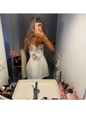 Vit kort klänning med blommig rosett - Supersöt vit kort klänning med en stor rosett framtill i blommigt mönster. Axelbandslös modell med markerad midja och utsvängd kjol. Perfekt för sommarens alla tillfällen.