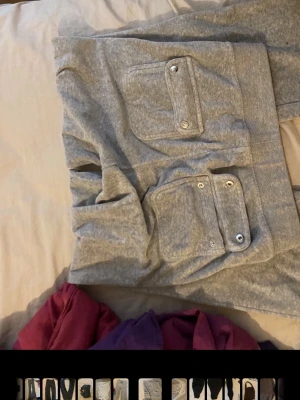Gråa juicy couture byxor - Säljer ett par grå mjukisbyxor med raka ben och stora fickor framtill med lock och tryckknappar. Byxorna har dragsko i midjan och är gjorda i mjukt, bekvämt material. Perfekta för chill dagar hemma eller när du vill vara extra comfy.