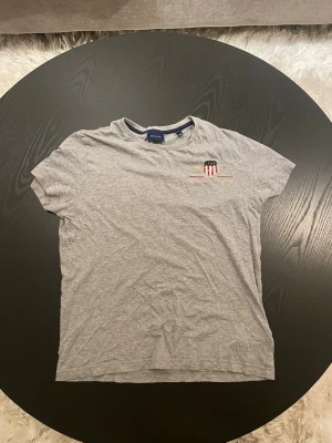 Grå t-shirt från Gant - Snygg grå t-shirt från Gant med ett litet tryck på bröstet i form av ett emblem med rött, vitt och blått. Klassisk passform och rund halsringning. Perfekt till jeans eller shorts. Storlek XS