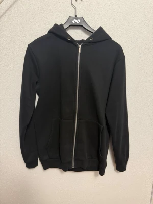 Svart zip hoodie från SMOG - En svart hoodie från SMOG i slim fit-modell med dragkedja framtill och två stora fickor. Klassisk huva och ribbade muddar vid ärmar och nederkant. Perfekt för en chill och enkel stil. Materialet känns mjukt och bekvämt.
