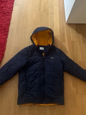 Svart pufferjacka med huva - Snygg Marinblå pufferjacka med gul insida och huva. Jackan har dragkedja framtill, två sidofickor och quiltad design. Perfekt för kalla vinterdagar och ger en sportig look. Liten logga på bröstet för extra stilpoäng.