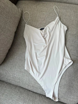 Ljusbeige body med tunna axelband - Säljer en ljusbeige body från Trendyol med tunna axelband och draperad ringning. Materialet är mjukt och stretchigt, vilket ger en skön passform. Perfekt att styla till olika outfits för en clean och fräsch look.