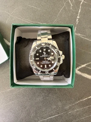 Rolex Submariner silverklocka - Stilren Rolex Submariner med svart urtavla och silverfärgad länk i metall. Klockan har tydliga markeringar, datumvisning och klassisk vridbar bezel. Perfekt accessoar för dig som gillar exklusiv och tidlös design.