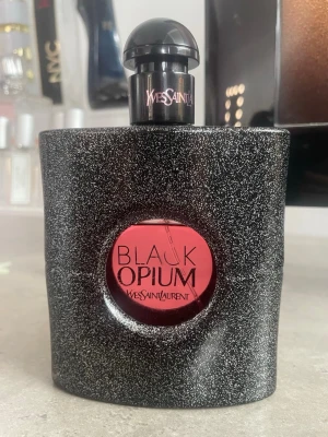 Yves Saint Laurent Black Opium parfym - Black Opium från Yves Saint Laurent är en ikonisk parfym i en svart, glittrig flaska med rund rosa detalj och svart kork. Flaskan har en modern och lyxig design som sticker ut på parfymhyllan. Perfekt för dig som gillar djärva och eleganta dofter.