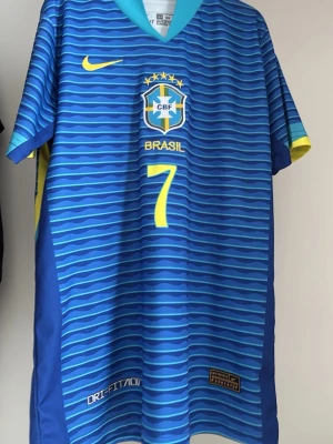 Brasilien bortaställ vini jr - Snygg blå Brasilien fotbollströja från Nike med gula detaljer och nummer 7 på både fram- och baksidan. På ryggen står det 'VINI JR.'. Tröjan har vågiga ränder, korta ärmar och är tillverkad i ett lätt, snabbtorkande material med Dri-FIT-teknologi.