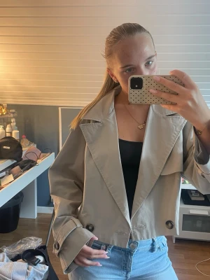 Kort beige trenchcoat  - Snygg kort trenchcoat. Jackan har dubbelknäppning med stora knappar och breda ärmslut med dekorativa spännen. Perfekt för en trendig och avslappnad look.