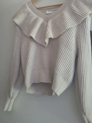 Beige stickad tröja med volang H&M - Supersnygg beige stickad tröja från H&M med bred volang runt v-ringningen. Tröjan har ribbstickat mönster, långa ärmar och något kortare framkant för en trendig look. Perfekt att matcha med jeans eller kjol.