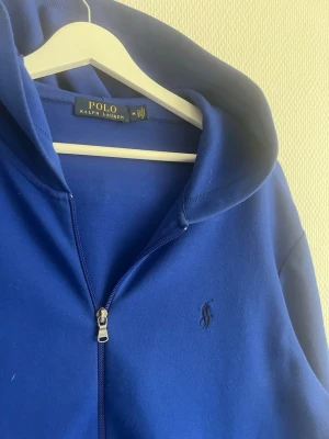 Blå hoodie från Polo Ralph Lauren - Snygg blå hoodie från Polo Ralph Lauren med dragkedja framtill och klassisk huva. Tröjan har två fickor på magen och den ikoniska broderade loggan på bröstet. Perfekt för en chill och stilren look. Materialet känns mjukt och bekvämt.