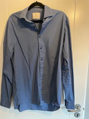 Blå skjorta från The Shirt Factory - Snygg blå skjorta från The Shirt Factory i slim fit-modell. Skjortan är lättstruken och har klassisk krage, långa ärmar och knappar framtill. Perfekt för dig som vill ha en stilren och enkel look.