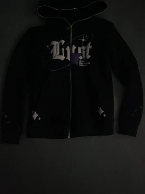 Svart hoodie med tryck och dragkedja - Säljer en svart hoodie med huva och dragkedja framtill. På bröstet finns ett stort tryck i vitt och lila med texten 'Lust' i gotisk stil. Hoodien har fickor på sidorna och detaljer i vitt. Perfekt för en streetwear-look.