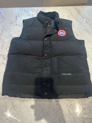 Canada Goose väst - Snygg svart dunväst från Canada Goose med hög krage och tryckknappar framtill. Västen har två stora fickor och den klassiska Canada Goose-loggan på bröstet. Perfekt för lager-på-lager och håller dig varm under kyliga dagar.