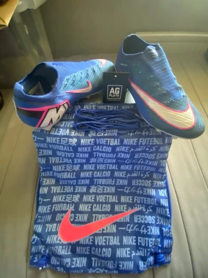 Nike Mercurial Vapor 16 Elite AG Stl 42 - Säljer ett par Nike Mercurial fotbollsskor i blått med vita och rosa detaljer. Skorna har en strumpliknande passform och är tillverkade i ett lätt, ventilerande material. Medföljer blå skopåse med Nike-logga och extra skosnören. Perfekta för konstgräs.