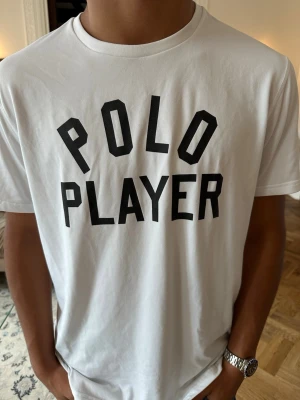 Vit polo t-shirt med svart tryck - Vit t-shirt med texten 'POLO PLAYER' i svart på bröstet. Klassisk rund halsringning och korta ärmar. Tillverkad i mjukt bomullstyg som känns skönt mot huden. Perfekt för en avslappnad och stilren look.
