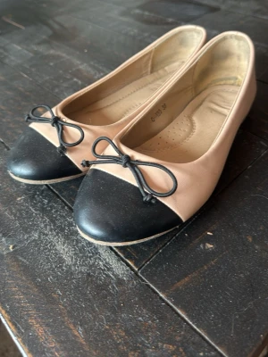Beige och svarta ballerinaskor med rosett - Snygga ballerinaskor i beige med svart tåparti och dekorativa svarta rosetter framtill. Skorna har rundad tå och låg sula, perfekta för en stilren och klassisk look. Tillverkade i ett slätt material och är väldigt sköna. 