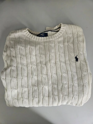 Vit kabelstickad tröja Polo Ralph Lauren - Kabelstickad tröja i offwhite från Polo Ralph Lauren. Klassisk rund halsringning och diskret broderad logga på bröstet. Tröjan har ribbade muddar och är gjord i mjukt material, perfekt för lager på lager under kyliga dagar. (Kids M) köpt för 1099 sek