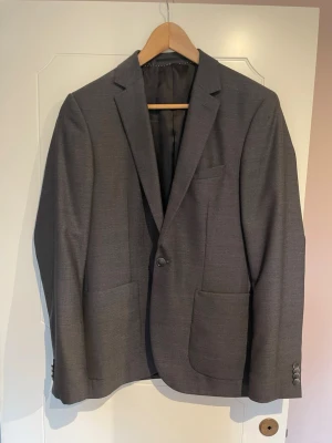 Mörkgrå kavaj, slim fit strl 48 - Stilren mörkgrå kavaj med två framfickor och en bröstficka. Kavajen har smal passform, enkelknäppt design och klassiska. Perfekt för dig som vill ha en clean och tidlös look.