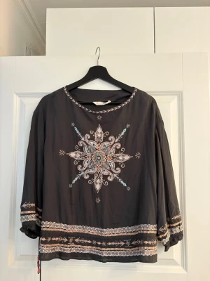 Blus Odd Molly - Mörkgrå blus från Odd Molly med vackra broderier i vitt, rosa och turkost framtill och längs ärmslut och nederkant. Blusen har rund halsringning och långa, lösa ärmar med resår vid ärmslut. 