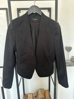 Svart kavaj från Lindex - Stilren svart kavaj från Lindex med öppen front och figurnära passform. Kavajen har långa ärmar, två fickor framtill och är helfodrad. Perfekt för en snygg och klassisk look.