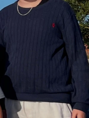 Mörkblå kabelstickad tröja Polo Ralph Lauren - Mörkblå kabelstickad tröja från Polo Ralph Lauren med rund halsringning och röd broderad logga på bröstet. Tröjan har långa ärmar och ribbade muddar vid ärmslut och nederkant. Klassisk och stilren modell i mjukt material.