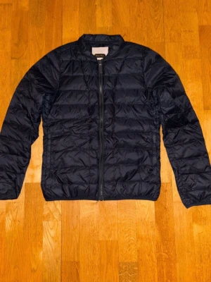 Svart pufferjacka med dragkedja - Snygg svart pufferjacka med quiltad design och rak passform. Jackan har en hög krage och hel dragkedja framtill. Tillverkad i ett glansigt, lätt material som håller värmen under kalla dagar. Perfekt för dig som vill ha en stilren och enkel vinterjacka.