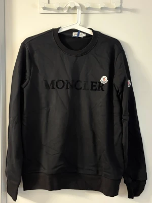 Svart sweatshirt från Moncler - Stilren svart sweatshirt från Moncler med broderad logga på bröstet och ärmen. Tröjan har rund halsringning, ribbade muddar och en klassisk passform. Perfekt för dig som gillar exklusiva streetwear-plagg och vill ha något som sticker ut.