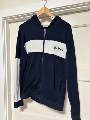 Marinblå hoodie med dragkedja från BOSS - Snygg marinblå hoodie från BOSS med vit bred rand över bröstet och på ärmarna. Dragkedja framtill, två fickor och huva. BOSS-logga tryckt på bröstet. Perfekt för en avslappnad och stilren look.
