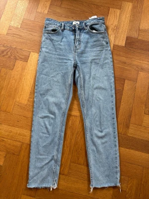 Blå raka jeans från ONLY - Säljer ett par ljusblå raka jeans från ONLY med råa, fransiga benslut. Klassisk femficksmodell med dragkedja och knapp. Jeansen har en avslappnad passform och är tillverkade i denim med synliga sömmar.