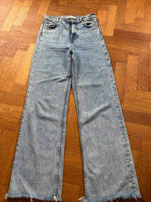 Vida ljusblå jeans från ZARA - Säljer ett par ljusblå jeans från ZARA med hög midja och vida ben. Jeansen har råa, fransiga benslut och klassisk femficksdesign. Tillverkade i denim med dragkedjegylf och knapp. Perfekta för en avslappnad och trendig look.