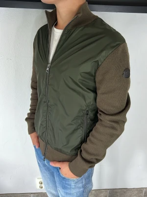 Moncler Cardigan  - Snygg olivgrön bomberjacka med stickade ärmar och rygg i mörkare grön ton. Jackan har dragkedja framtill, ribbad krage och muddar samt två sidofickor med dragkedja. Liten patch på ärmen. Perfekt för dig som vill ha en modern och avslappnad stil. Large passar M