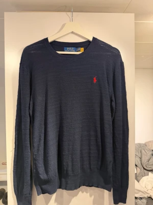Mörkblå stickad tröja Polo Ralph Lauren - Mörkblå stickad tröja från Polo Ralph Lauren med rund halsringning och röd broderad logga på bröstet. Långärmad modell med ribbade muddar vid ärmslut och nederkant. Perfekt för lager på lager och stilren look. Helt oanvänd 