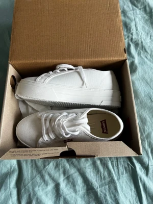 Vita Levi's Tijuana sneakers med platåsula - Snygga helvita sneakers från Levi's med kraftig platåsula och klassisk låg design. Skorna har ovandel i canvas, vita skosnören och en liten röd Levi's-tag på sidan. Perfekta för dig som vill ha en clean och stilren look till vardagsoutfiten.