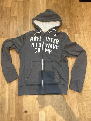 Grå Hollister zip hoodie med tryck - Grå hoodie från Hollister med vit dragkedja och trycket 'Hollister Big Wave Comp.' på bröstet. Mjuk insida, två fickor fram och justerbar huva med vita snören. Perfekt för chill dagar och enkelt att matcha 