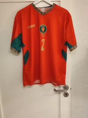 Marocko fotbollströja Hakimi #2 - Säljer en röd Marocko fotbollströja med gröna detaljer och guldfärgade tryck. Tröjan har nummer 2 och Hakimi på ryggen, samt Marockos emblem på bröstet. Unik mönstrad kant vid hals och ärmslut. Perfekt för dig som älskar fotboll och vill stötta Marocko.