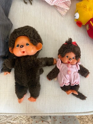 Två Monchhichi apdockor - Säljer två söta Monchhichi apdockor med mjuk, brun päls och plastansikten. Den ena har en helbrun dräkt och den andra har en rosa overall med rosett i håret. Perfekta för samlare eller som present!