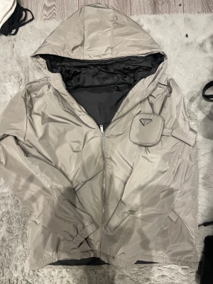 Beige vindjacka från Prada med huva - Snygg beige vindjacka från Prada med huva och dragkedja framtill. Jackan har en liten ficka med lock och Prada-logga på bröstet samt elastiska muddar vid ärmsluten. Tillverkad i glansigt nylonmaterial och helsvart innerfoder. Perfekt för en trendig streetwear-look.