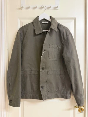 Olivgrön overshirt med fickor - Stilren olivgrön overshirt i kraftigt bomullstyg. Klassisk rak passform med tre utanpåliggande fickor framtill och knäppning med stora knappar. Perfekt lager-på-lager-plagg med enkel krage och diskreta sömmar.
