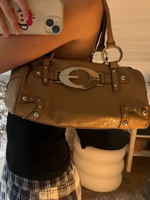 Beige guess väska med metallspänne - Säljer en beige handväska i skinnimitation med stora silverfärgade metalldetaljer och nitar. Väskan har ett ovalt metallspänne framtill och dubbla handtag i samma färg. Cool och rymlig modell med dragkedja och dekorativa sömmar.
