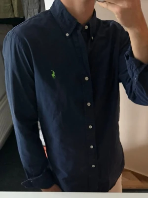 Mörkblå skjorta från Ralph Lauren - Klassisk mörkblå skjorta från Ralph Lauren med button-down krage och grönt broderat logotyp på bröstet. Skjortan har långa ärmar, vita knappar och är tillverkad i bomull. Perfekt för en clean och stilren look.