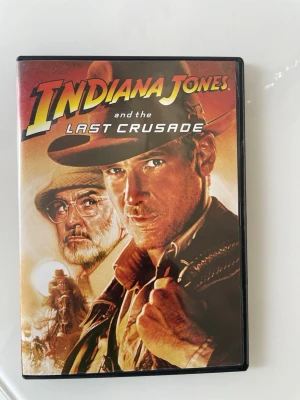Indiana Jones and the Last Crusade DVD - DVD-film med omslag till Indiana Jones and the Last Crusade. Omslaget har en illustration av Indiana Jones och hans far, samt äventyrstema med varma färger. Text och bilder på baksidan beskriver filmens handling och extramaterial.