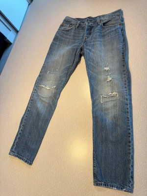 501 Levi's jeans blå slitna - Säljer ett par klassiska blå Levi's jeans 501 med slitningar och hål på benen för en cool, avslappnad look. Jeansen har fem fickor, raka ben och den ikoniska läderlappen bak i midjan. Perfekta för dig som gillar vintage vibe och streetstyle.