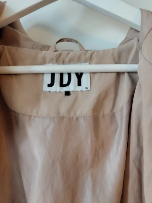 Beige vindjacka från JDY med huva - Snygg beige vindjacka från JDY i storlek XL. Jackan har huva, dragkedja och tryckknappar framtill samt två fickor med lock. Dragsko i midjan för justerbar passform. Perfekt för blåsiga dagar och enkel att matcha med olika outfits.