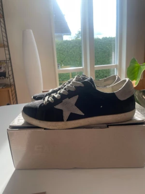 Svarta sneakers med stjärndetalj - Säljer ett par svarta sneakers med vit stjärna på sidan och vita skosnören. Skorna har en låg profil och vit sula. Materialet ser ut att vara mocka och textil, vilket ger en cool och avslappnad look.