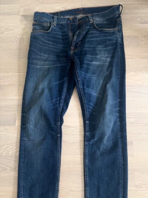 Mörkblå raka jeans från Nudie - Säljer ett par mörkblå jeans från Nudie CO med raka ben och klassisk femficksmodell. Jeansen har snygga slitningar och kontrastsömmar samt patch med logga bak i midjan. Tillverkade i denim av bomull för en tidlös look. Storlek 34/32. 